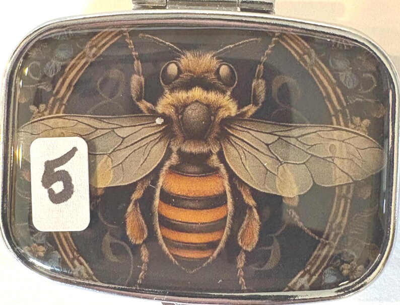 Puede incluir: Un pastillero rectangular plateado con una ilustraci&oacute;n detallada de una abeja. La abeja es marr&oacute;n y naranja, con alas transparentes. El n&uacute;mero "5" est&aacute; en una pegatina blanca en la esquina inferior izquierda. El fondo es oscuro con detalles florales.