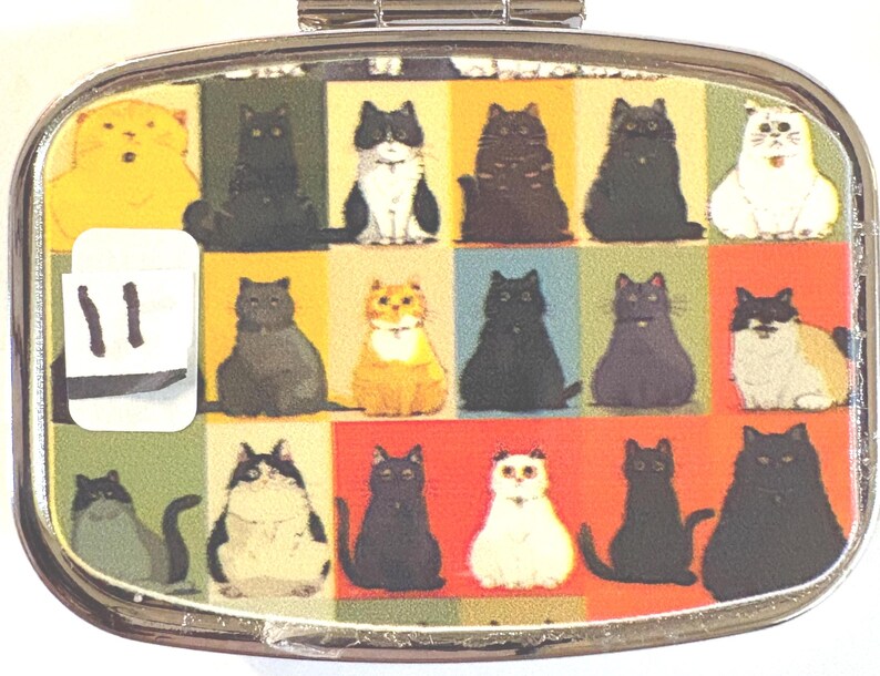 May include: A silver-toned, oval-shaped pill case with a colorful cat-themed design. The case features a grid of various cat illustrations in different colors and poses. A white sticker with the number "11" is on the left side.