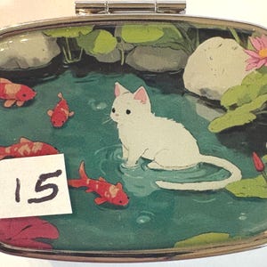 May include: A rectangular, silver-edged pill box with a painted scene of a white cat in a pond with koi fish and lily pads. The number "15" is on a white sticker in the lower left corner.