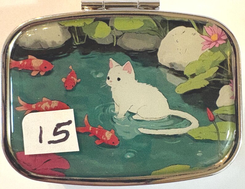 May include: A rectangular, silver-edged pill box with a painted scene of a white cat in a pond with koi fish and lily pads. The number "15" is on a white sticker in the lower left corner.