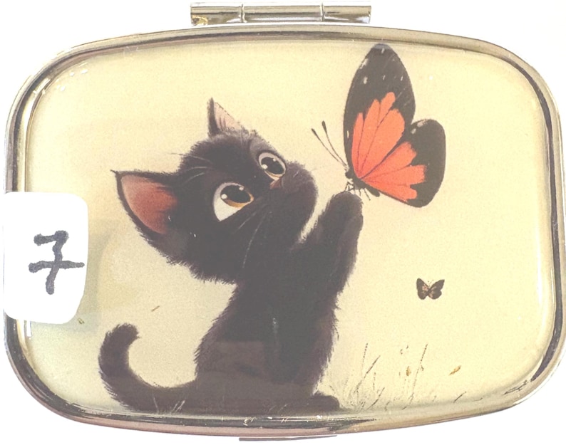 May include: A rectangular, hinged pill box with a cartoon illustration of a black kitten reaching for a butterfly with orange and black wings. The background is a light yellow color. The number 7 is on a white sticker in the corner.