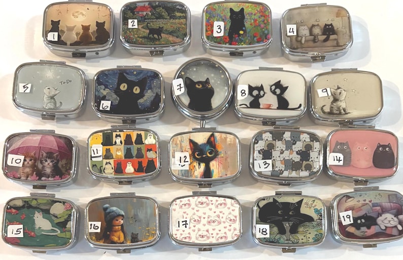 May include: A collection of rectangular, silver-toned pill boxes, each featuring a unique cat-themed design. The boxes showcase various cat illustrations, including black cats, kittens under an umbrella, and cat patterns. The designs are colorful and artistic.
