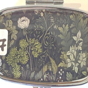 Puede incluir: Un pastillero rectangular plateado con un dise&ntilde;o floral. El interior presenta un fondo oscuro con un patr&oacute;n de flores y follaje verdes y crema. Una pegatina blanca con el n&uacute;mero "17" es visible.