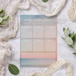 May include: A weekly schedule planner with a watercolour background of a sunset over the ocean. The planner has spaces for each day of the week, Monday through Sunday, and a notes section.