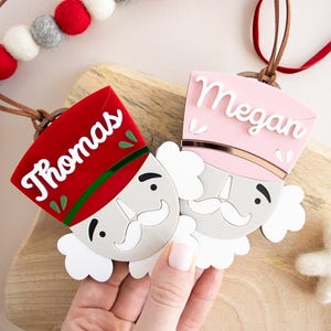Könnte beinhalten: Zwei personalisierte Weihnachtsschmuckstücke in Form von Spielzeugsoldaten. Eines ist rot mit "Thomas" in Weiß, das andere ist rosa mit "Megan" in Weiß. Beide haben einen weißen Schnurrbart und eine braune Lederschnur.