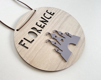 Pode incluir: Uma placa redonda de madeira com o nome "FLORENCE" recortado em letras grandes e em negrito. Uma silhueta de castelo cinza está à direita. Um cordão de couro marrom é preso para pendurar. A placa tem um acabamento de grão de madeira clara.