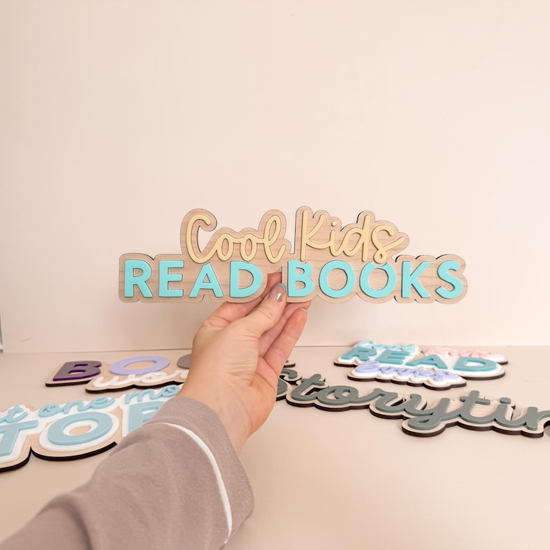 May include: Wooden sign with the text "Cool Kids READ BOOKS" in light blue and beige. The sign is held by a person. Other wooden signs with text are visible in the background.
