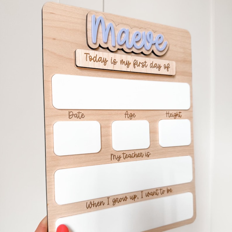 Op de afbeelding: Houten bord voor de eerste schooldag voor Maeve, met lichtblauwe letters. Het bord bevat vakken om de datum, leeftijd en lengte in te vullen. De tekst op het bord luidt "Vandaag is mijn eerste schooldag", "Mijn leraar is" en "Als ik groot ben, wil ik zijn".