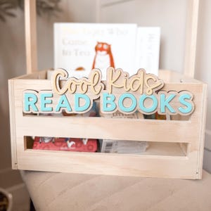 May include: A light-colored wooden crate with a handle, holding books. The crate has the words "Cool Kids READ BOOKS" in layered, light brown and turquoise letters. The crate is filled with books, including one with a tiger illustration.