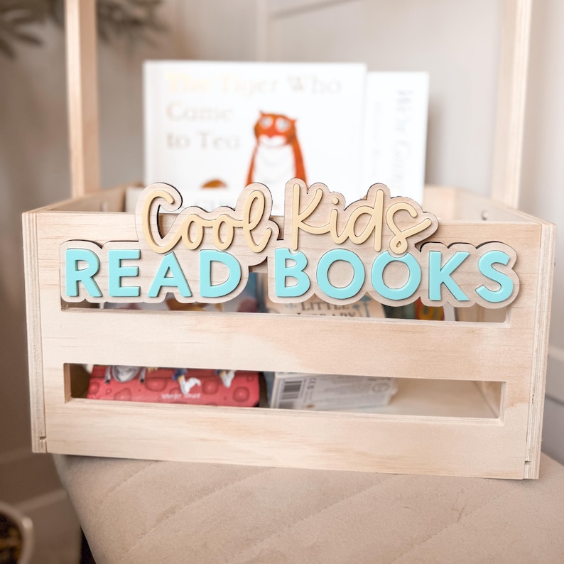 May include: A light-colored wooden crate with a handle, holding books. The crate has the words "Cool Kids READ BOOKS" in layered, light brown and turquoise letters. The crate is filled with books, including one with a tiger illustration.
