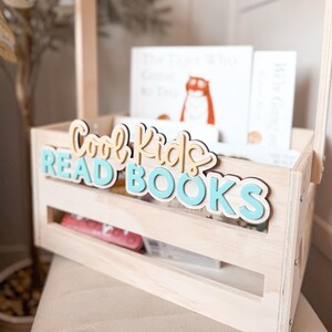 May include: A light wooden crate with a handle, filled with books. The crate has the words "Cool Kids READ BOOKS" in layered lettering. The books inside have white covers and colorful illustrations. A perfect storage solution for children's books.