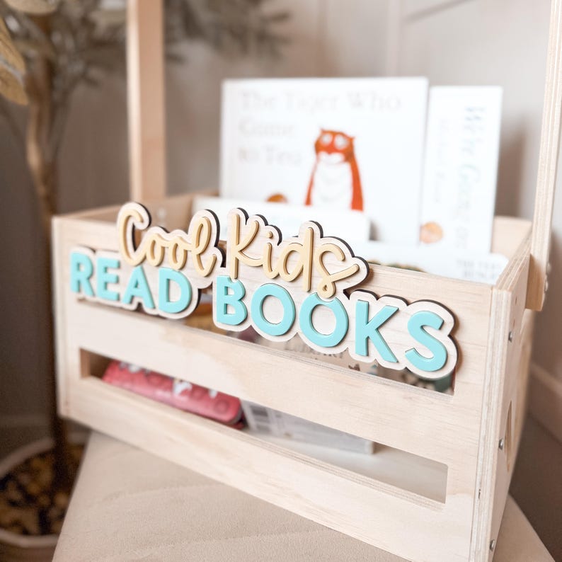 May include: A light wooden crate with a handle, filled with books. The crate has the words "Cool Kids READ BOOKS" in layered lettering. The books inside have white covers and colorful illustrations. A perfect storage solution for children's books.