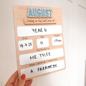 Op de afbeelding: Een houten bord met het woord "AUGUST" in blauwe letters. Het bord bevat velden voor "Jaar 6", "Datum", "Leeftijd", "Lengte", "Mijn leraar is" en "Als ik groot ben, wil ik zijn". De lengte is 119 cm.