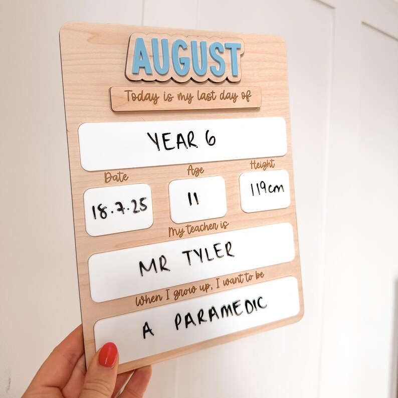 Op de afbeelding: Een houten bord met het woord "AUGUST" in blauwe letters. Het bord bevat velden voor "Jaar 6", "Datum", "Leeftijd", "Lengte", "Mijn leraar is" en "Als ik groot ben, wil ik zijn". De lengte is 119 cm.