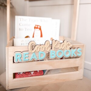 May include: A wooden crate with the words "Cool Kids READ BOOKS" in light blue and tan lettering. The crate holds several books, including one titled "The Tiger Who Came to Tea." The crate is made of light-colored wood.
