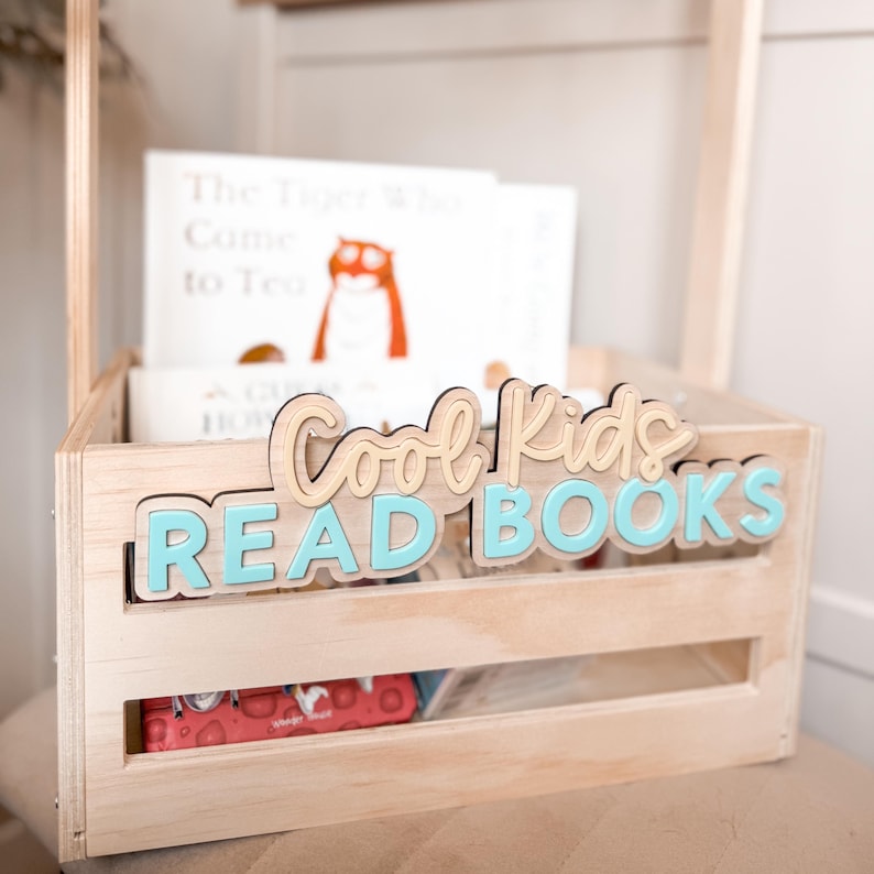 May include: A wooden crate with the words "Cool Kids READ BOOKS" in light blue and tan lettering. The crate holds several books, including one titled "The Tiger Who Came to Tea." The crate is made of light-colored wood.
