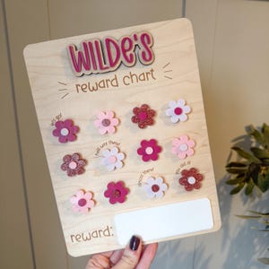 May include: Wooden reward chart with the name "WILDE'S" in pink lettering. The chart features flower-shaped tokens in various shades of pink and a space for rewards. Text includes "reward chart", "lets go!", "half way there!", "almost there!", and "you did it!"