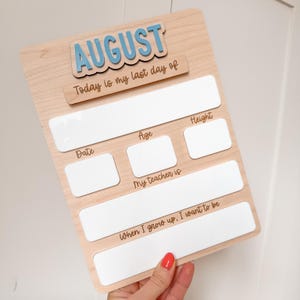 Op de afbeelding: Houten bord met het woord "AUGUST" in blauw en de tekst "Today is my last day of". Het bord heeft vakken voor "Date", "Age", "Height", "My teacher is" en "When I grow up, I want to be".