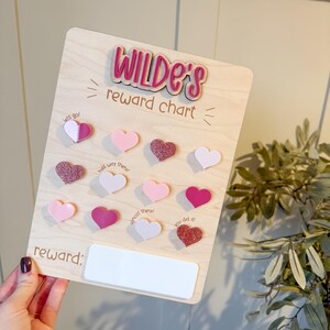 May include: A wooden reward chart with the name "Wilde's" in pink lettering. The chart has pink heart-shaped tokens and phrases such as "lets go!" and "you did it!" A white rectangle at the bottom is labelled "reward:"