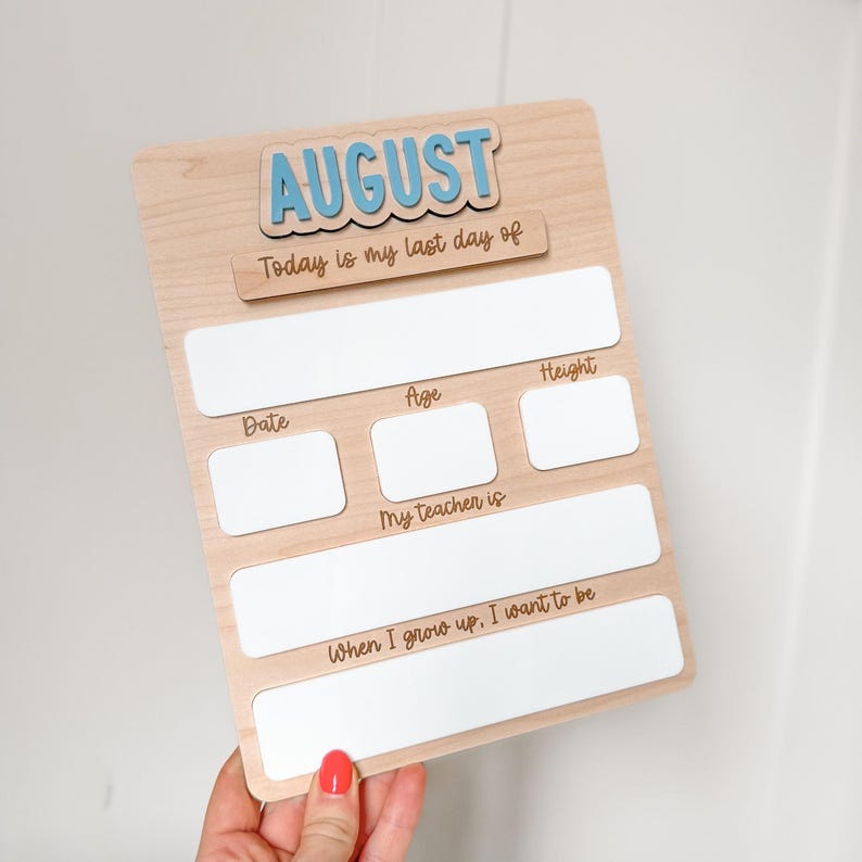 Op de afbeelding: Houten bord met het woord "AUGUSTUS" in blauwe letters. Het bord bevat vakken om de datum, leeftijd, lengte, naam van de leraar en toekomstige ambities in te vullen. De tekst "Today is my last day of" is ook opgenomen.