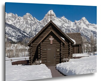 May include: A dark brown log chapel with a cross on the roof peak and a wooden door, situated in a snowy landscape. Behind the building, jagged snow-covered mountain peaks rise against a clear blue sky. Small signs are visible near the entrance.