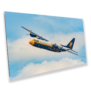 May include: A photograph of a blue and yellow C-130 Hercules aircraft, with the words "Blue Angels" and "United States Navy" visible. The aircraft is in flight against a backdrop of a cloudy blue sky.