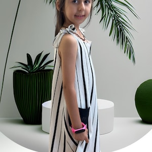 以下が含まれることがあります： 白い壁の前で、白と黒のストライプのノースリーブドレスを着た若い女の子が、ピンクの腕時計をしています。彼女の隣には、鉢植えの植物と緑色のボールがあります。