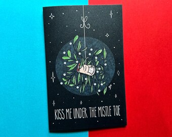 May include: A black card with a white illustration of a hand under a sprig of mistletoe. The text "Kiss me under the mistletoe" is printed in white at the bottom of the card.