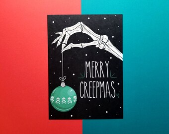 May include: A black card with a white skeleton hand holding a green Christmas ornament with skull and crossbones. The card says "Merry Creepmas".