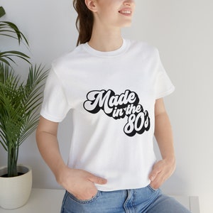 以下が含まれることがあります： レトロなフォントで「Made in the 80's」と書かれた黒いテキストがプリントされた白いTシャツです。