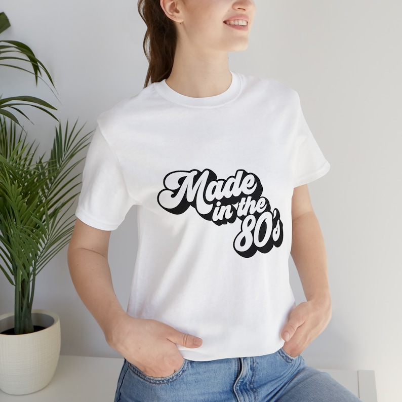 以下が含まれることがあります： レトロなフォントで「Made in the 80's」と書かれた黒いテキストがプリントされた白いTシャツです。