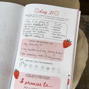 K&ouml;nnte beinhalten: Ge&ouml;ffnete Tagebuchseite mit handschriftlichen Notizen und Aufforderungen zur Selbstreflexion. Die Seite enth&auml;lt die &Uuml;berschrift "Tag 21" und Abschnitte f&uuml;r ein Brain Dump, eine To-Do-Liste und Dankbarkeit. Dekorative Erdbeer-Illustrationen sind vorhanden.