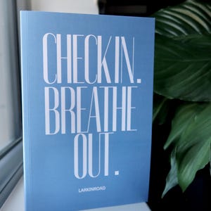 K&ouml;nnte beinhalten: Ein hellblaues Buch mit den Worten "CHECKIN. BREATHE OUT." in wei&szlig;er, stilisierter Schrift. Der Text "LARKINROAD" ist darunter gedruckt. Das Buch liegt auf einer wei&szlig;en Oberfl&auml;che, mit einer gr&uuml;nen Pflanze im Hintergrund.