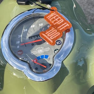 May include: An orange glittery keychain with the words "KEEP IT 100" hangs from a scooter's dashboard. The scooter's speedometer shows a speed of 20 mph. The dashboard is green with a chrome bezel.