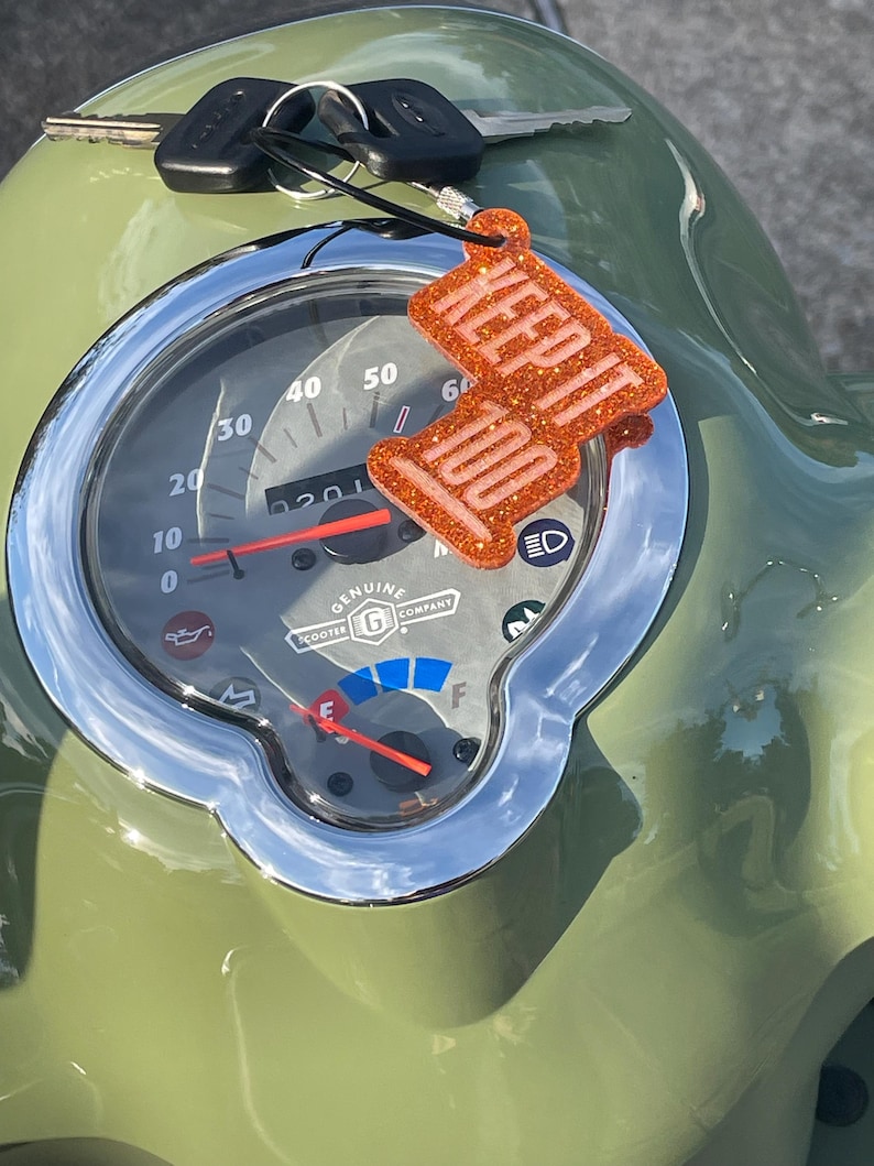 May include: An orange glittery keychain with the words "KEEP IT 100" hangs from a scooter's dashboard. The scooter's speedometer shows a speed of 20 mph. The dashboard is green with a chrome bezel.