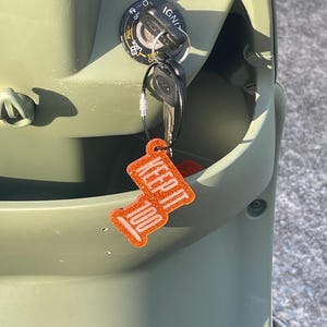May include: Close-up of a scooter's ignition with keys inserted. The keys have a bright orange keychain that reads "KEEP IT 100" in white lettering. The scooter is olive green.