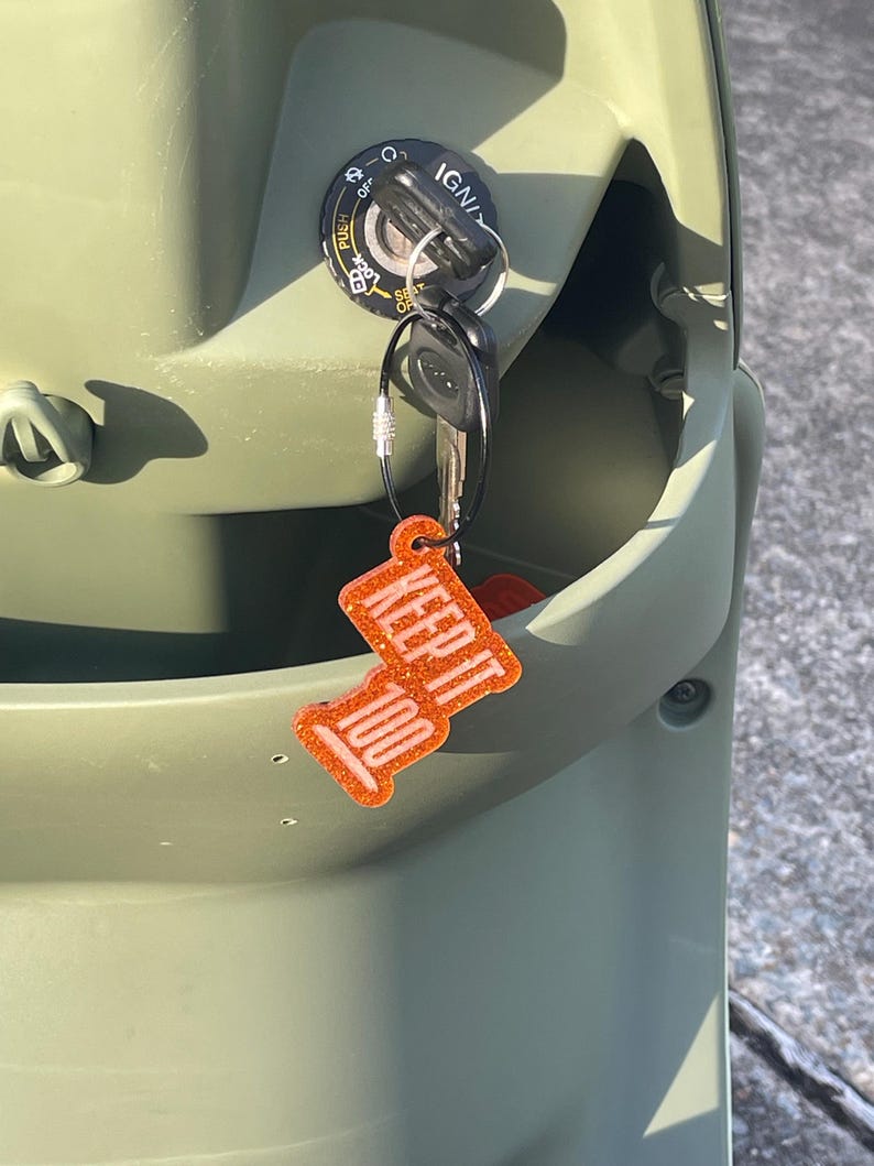 May include: Close-up of a scooter's ignition with keys inserted. The keys have a bright orange keychain that reads "KEEP IT 100" in white lettering. The scooter is olive green.
