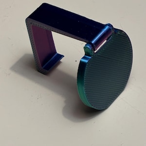 Peut inclure: Un support de téléphone imprimé en 3D, bleu et vert irisé. Le support a un dessus rectangulaire et une base incurvée et circulaire. L'intérieur du support est violet. Conçu pour maintenir un téléphone.