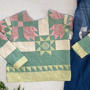 May include: A quilted top with a patchwork design in shades of green, pink, and cream. The long-sleeved shirt features a starburst pattern and a geometric border. Denim jeans are visible on the right side of the image.