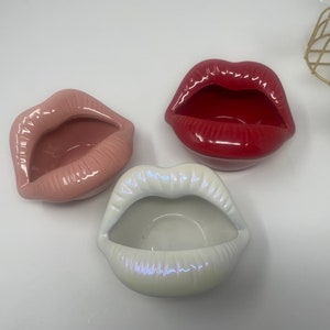 May include: Three ceramic ashtrays shaped like lips. One is pink, one is red, and one is white.
