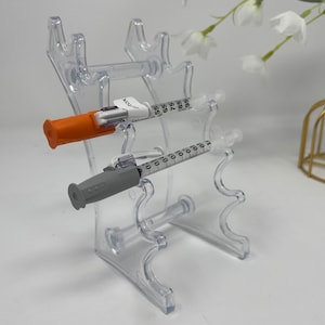 May include: Two clear plastic insulin syringes with orange and gray caps are displayed on a clear plastic stand. The stand has multiple slots for holding syringes.