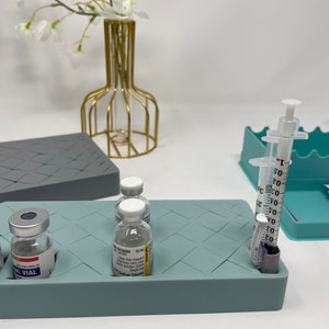 Puede incluir: Una bandeja de silicona azul claro con un patr&oacute;n de cuadr&iacute;cula, que contiene tres viales de vidrio transparentes etiquetados con "10 mL" y "VIAL", y dos jeringas de pl&aacute;stico transparentes con marcas en mililitros. La bandeja est&aacute; sobre una superficie blanca.