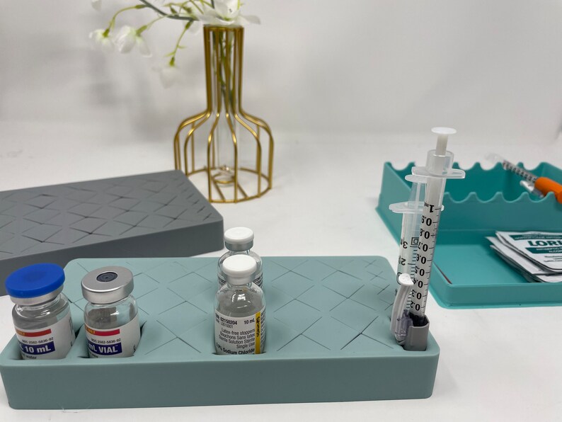Puede incluir: Una bandeja de silicona azul claro con un patr&oacute;n de cuadr&iacute;cula, que contiene tres viales de vidrio transparentes etiquetados con "10 mL" y "VIAL", y dos jeringas de pl&aacute;stico transparentes con marcas en mililitros. La bandeja est&aacute; sobre una superficie blanca.