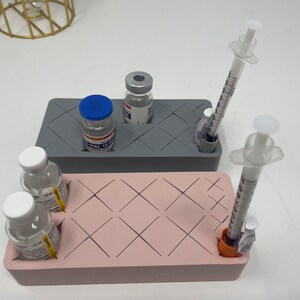 Puede incluir: Un organizador de silicona gris y rosa con compartimentos para viales y jeringas. El organizador tiene un patr&oacute;n de cuadr&iacute;cula y est&aacute; dise&ntilde;ado para contener suministros m&eacute;dicos.