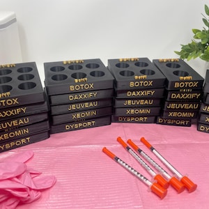 Op de afbeelding: Zwarte kunststof opbergdozen met gouden belettering voor verschillende soorten injectables. De dozen zijn gestapeld en gelabeld met de namen van de injectables: Botox, Daxxify, Jeuveau, Xeomin en Dysport.