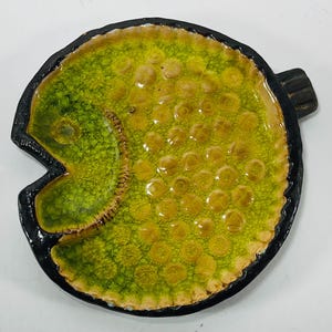 May include: A circular ceramic dish with a black rim and a vibrant lime green crackle glaze finish. The interior features a textured pattern of raised circular indentations across the surface, with a distinct crescent-shaped section on the left.