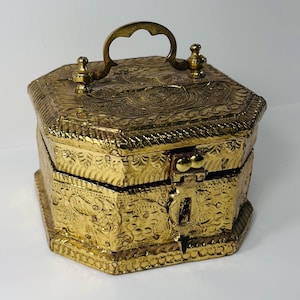May include: An octagonal brass box featuring intricate embossed patterns on all sides and the lid. The box includes a hinged lid with a central arched handle and a front latch mechanism designed for a lock, showcasing a metallic gold finish.