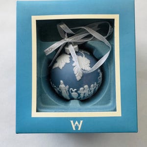 May include: A light blue spherical ornament with white relief figures and foliage designs, featuring a white ribbon with the text WEDGWOOD printed on it. The ornament is housed in a square blue box with a cream border and a white W logo at the bottom.