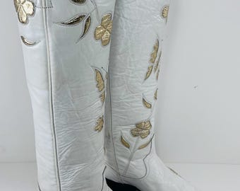 May include: A pair of white leather cowboy boots with gold floral and butterfly embroidery. The boots have a pointed toe and a black sole. The shaft of the boots reaches the knee.