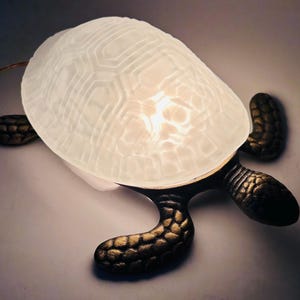 Pode incluir: Lâmpada decorativa em forma de tartaruga com casco branco texturizado e pernas e cabeça na cor bronze. A concha brilha com uma luz suave, criando um ambiente acolhedor. A lâmpada foi projetada para se assemelhar a uma tartaruga marinha.